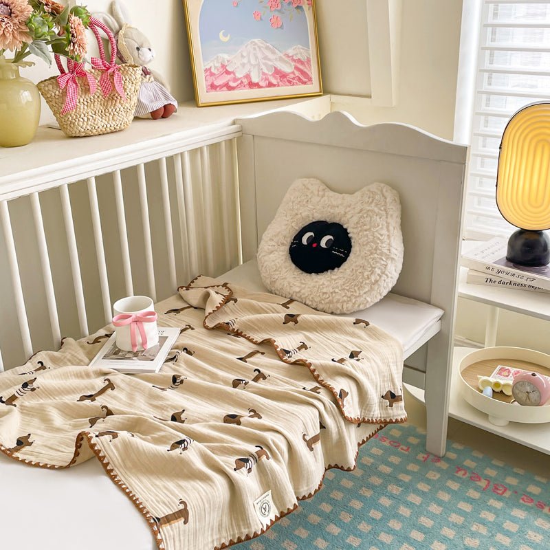 A beige textured wrinkled blanket with a cat pattern, draped over the edge of a bed next to a pillow with a cat face design. The room has a cozy decor with a framed picture on the wall, a basket, and a lamp.