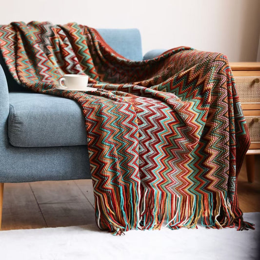 A colorful bohemian knit throw blanket with a geometric pattern and tassel trim, draped over the arm of a blue sofa.