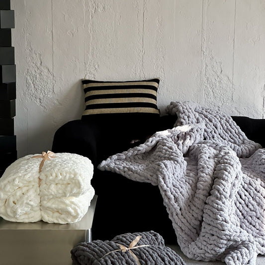A cozy chunky knit blanket in gray and white colors is draped over a armchair, with a cushion beside it. A rolled-up blanket with a bow on it is also visible in the foreground.