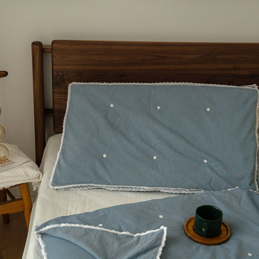 A blue silk pillow on a bed with a wooden headboard, the pillow has a solid pattern with what appears to be a natural fiber border.