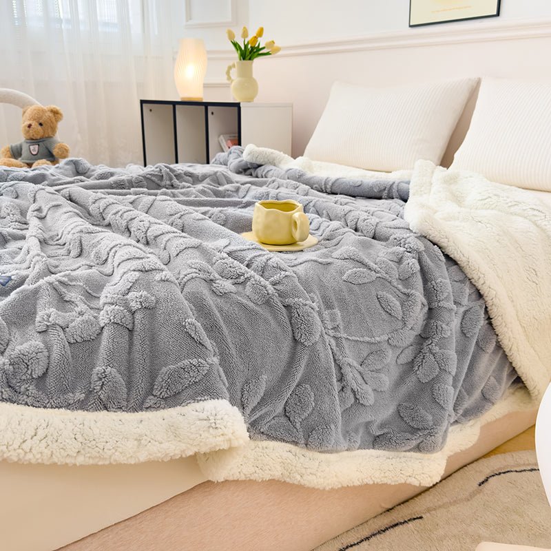 A gray solid sherpa throw with a floral pattern on a bed, with a white border. A teddy bear and a cup are visible on the bed.