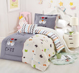 A children's bedding set with cat print, displayed on a bed in a room with white walls and furniture. The set includes a comforter, pillows, and a bedsheet with a cat and geometric design.