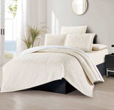 A beige bedspread displayed on a bed in a room with a plant and a mirror in the background.