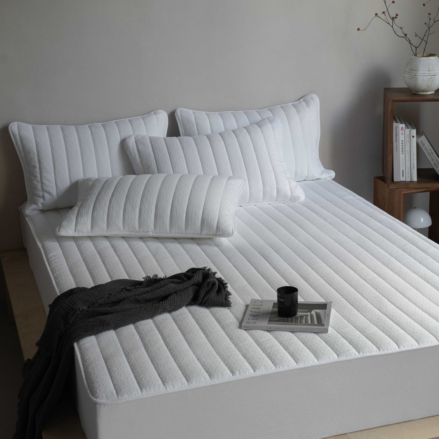 A white, waterproof, quilted fitted sheet on a bed, with several pillows on top and a black blanket draped over the side. A cup and a book are also visible on the bed.